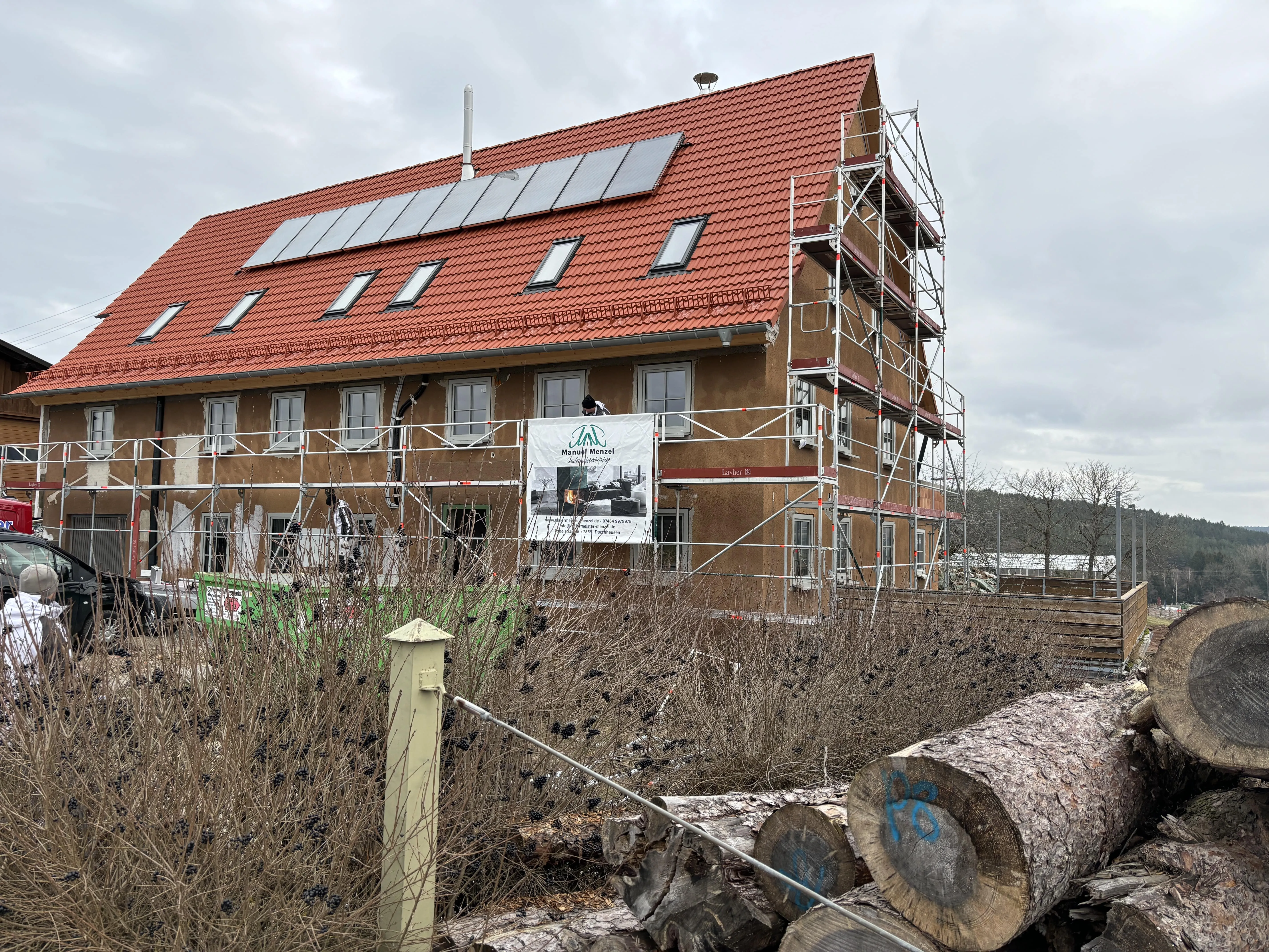 Eingerüstetes Wohnhaus mit rotem Ziegeldach und Solaranlage, im Vordergrund Holzstämme