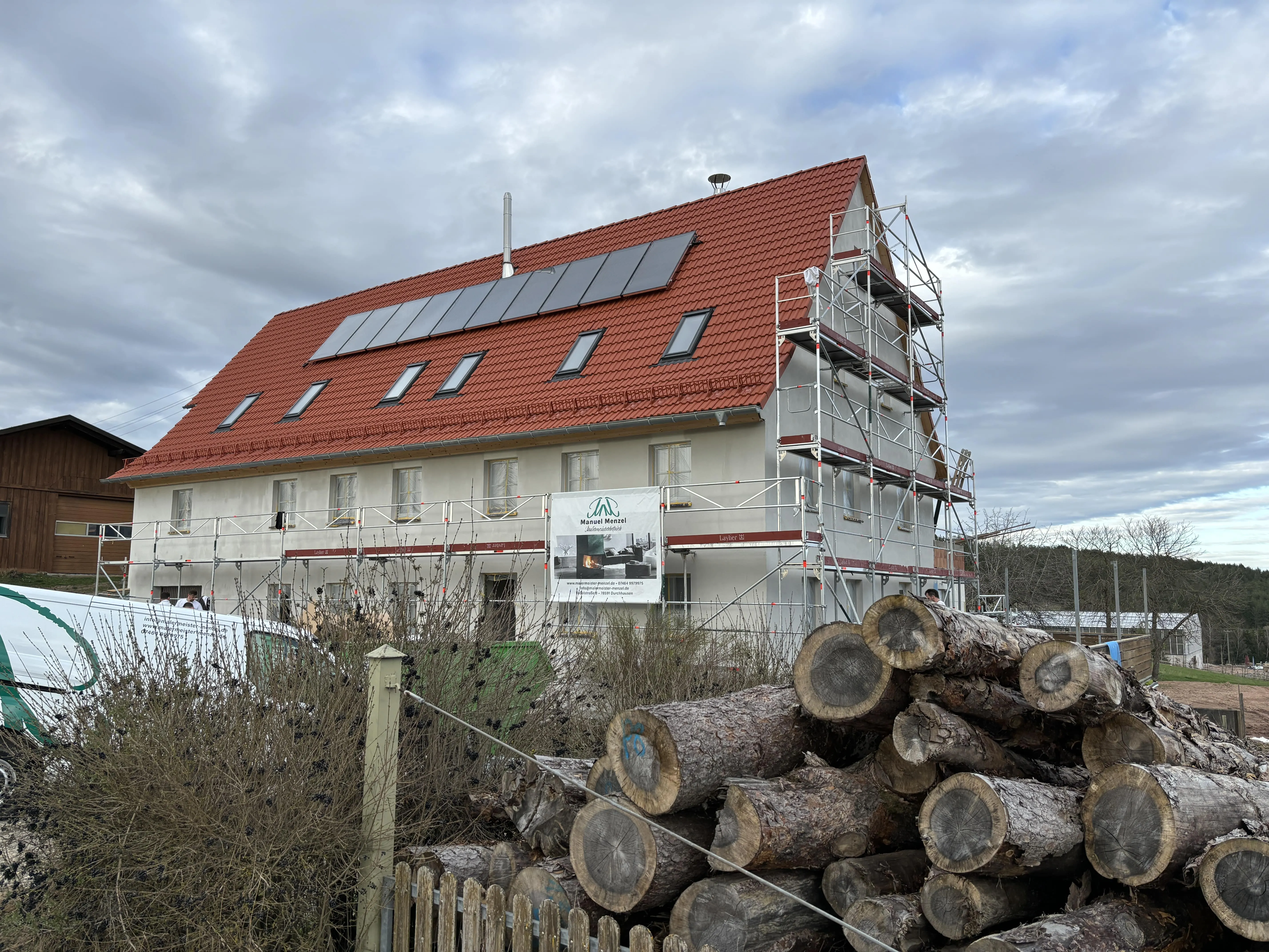 Wohnhaus mit Solardach und Gerüst an der Giebelseite, davor gestapelte Holzstämme